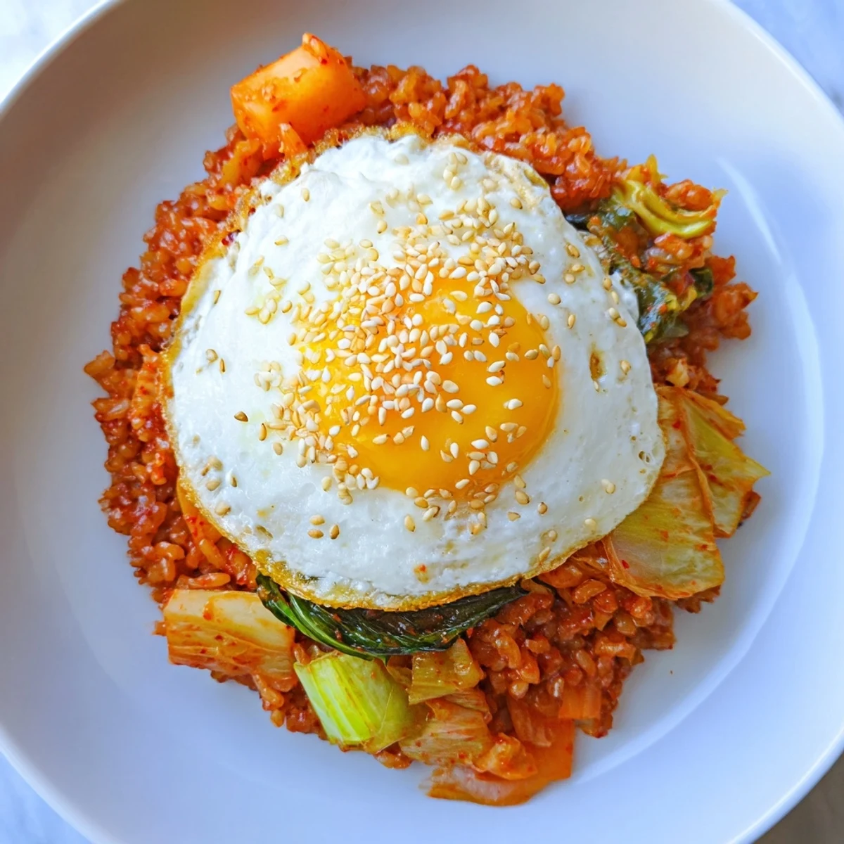A vibrant bowl of Kimchi Fried Rice topped with a golden fried egg and sesame seeds, next to a small dish of spicy kimchi.  