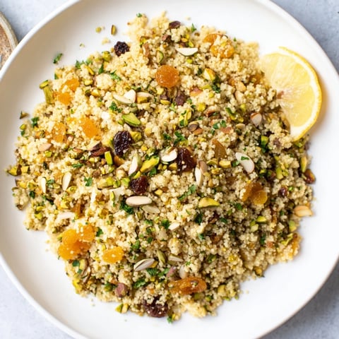 A close-up of fragrant couscous pilaf dotted with apricots, raisins, and colorful toasted nuts on a rustic plate.  