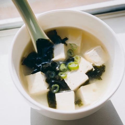 Silken tofu cubes and wakame seaweed swim in a steaming bowl of traditional miso soup, garnished with fresh scallions.