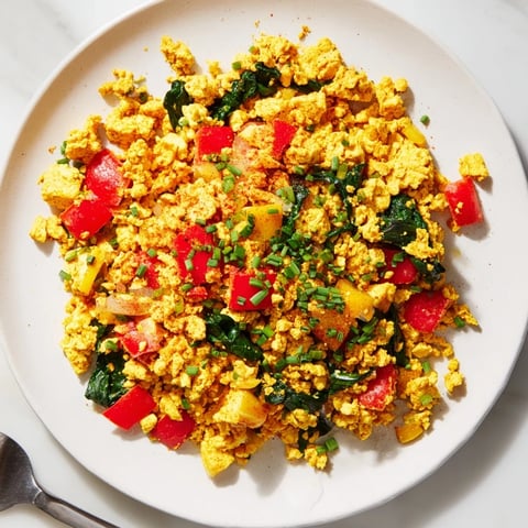 Golden tofu scramble sizzling with diced red bell pepper, onion, and fresh spinach in a skillet, topped with chopped chives and a dash of turmeric for a vibrant, plant-based breakfast.