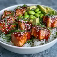 Golden-brown baked salmon cubes rest on fluffy jasmine rice, garnished with edamame, cucumber, and creamy avocado slices.